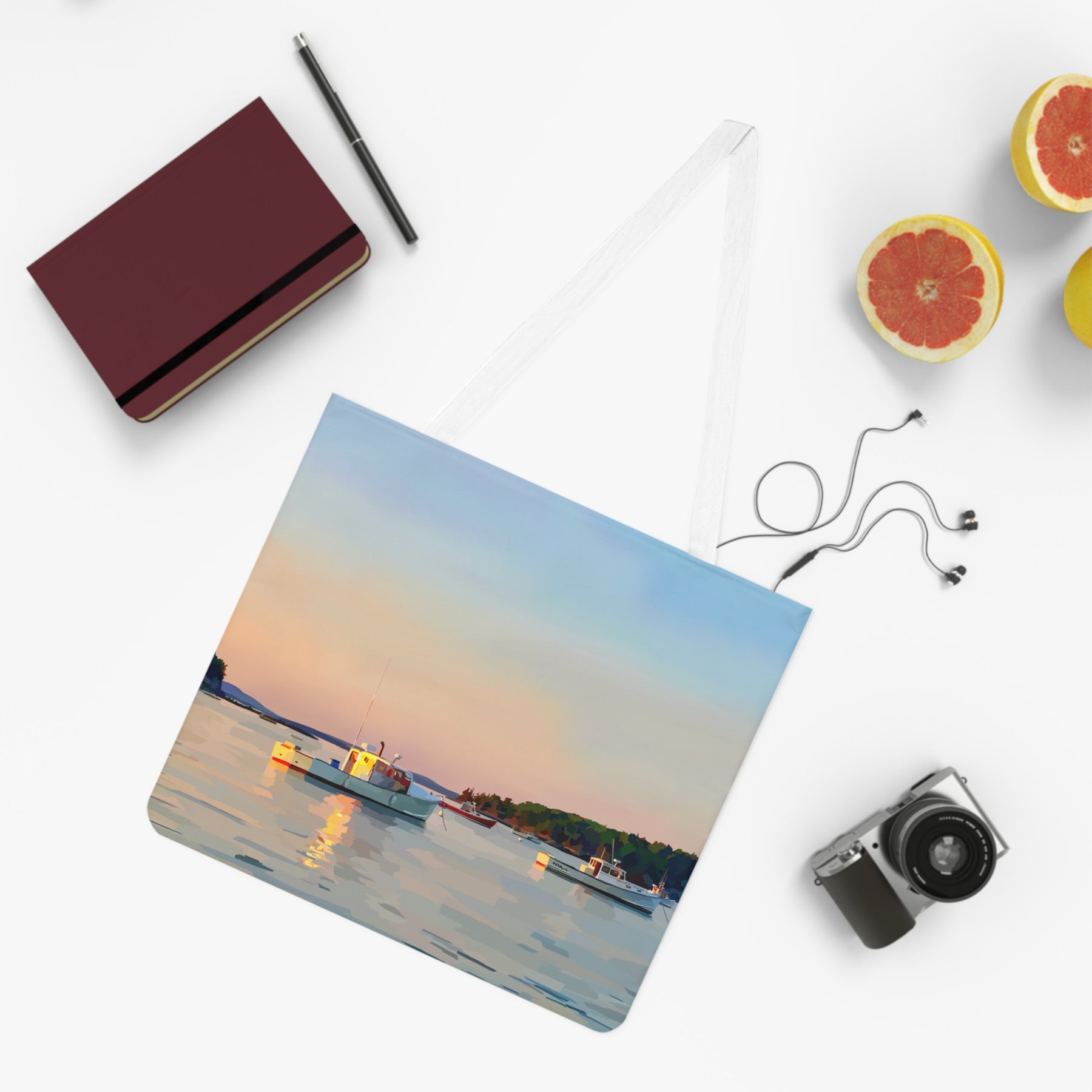 tote bag with a seascape painting of a sunset over the water with boats with a notebook, pen, camera, and fruit on a white surface